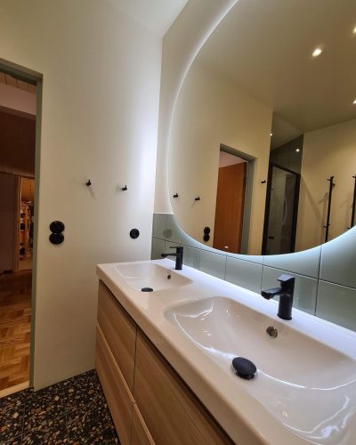 Modernt renoverat badrum med en stilren dubbel kommod i trä och en vit porslinstvättställ med svarta armaturer. Ovanför sitter en stor, rundad spegel med inbyggd belysning som skapar en elegant och mjuk ljuseffekt. Väggarna är delvis klädda i gröna kakelplattor och golvet har en terrazzo-yta med mörka och beige toner. Svarta detaljer som krokar och eluttag förstärker det minimalistiska uttrycket. Badrumsrenovering i Stockholm.