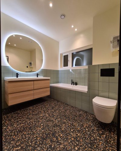 Stilrent renoverat badrum med grönkaklade väggar och ett golv i terrazzo med inslag av bruna och beige toner. Flytande dubbel kommod i trä med ett modernt rundat spegel med inbyggd belysning ovanför. Ett inbyggt badkar med svarta armaturer och vägghängd toalett skapar en minimalistisk och elegant känsla. Infälld takbelysning förstärker rummets atmosfär. Badrumsrenovering i Stockholm.
