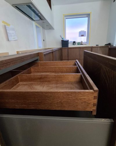 Köksrenovering i Stockholm med fokus på detaljer – en öppen låda i mörkt trä med elegant sinkfog visar hög hantverkskvalitet. Överskåp i vitt med integrerad fläkt och pågående arbete i bakgrunden.
