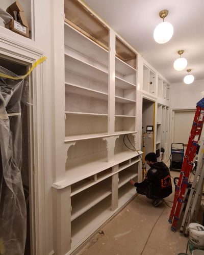 Pågående renovering av en platsbyggd vit bokhylla i en hall. En hantverkare från L&A Fantastic Renovering arbetar med hyllorna, och golvet är täckt med skyddspapper. Hallen har sekelskiftesdetaljer, mässingslampor och inbyggd förvaring. Finsnickeri och renovering i Stockholm.