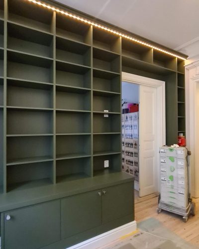 Platsbyggd grön bokhylla med LED-belysning, öppen förvaring och underskåp, integrerad runt en dörröppning i ett rum med sekelskiftesdetaljer. Pågående renovering med skyddspapper på golvet. Finsnickeri och renovering i Stockholm.