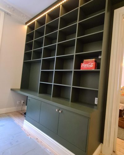 Platsbyggd grön bokhylla med öppen förvaring och skåp längst ned, installerad i ett rum med sekelskiftesdetaljer. LED-belysning längs toppen ger ett stilrent intryck. Finsnickeri och renovering i Stockholm.
