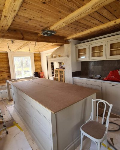 Pågående köksrenovering i ett rustikt hem med träpanel i tak och väggar. Ett stort köksö-skåp är installerat men saknar bänkskiva. Vita köksskåp med glasdörrar och en gammal murad spis bevaras i designen. Renovering i Stockholm.