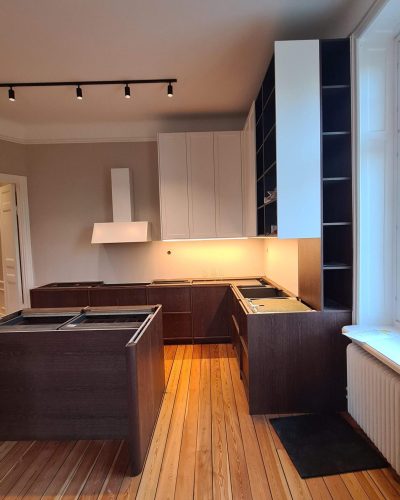 Pågående köksrenovering i Stockholm med ett modernt kök under installation. Köket har mörka träskåp, vita överskåp, en stilren vit fläkt och inbyggda hyllor. Trägolv och spotlights i taket skapar en varm atmosfär.