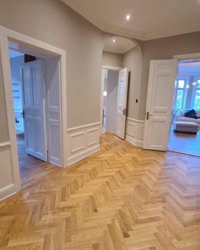 Nyrenoverad hall i en klassisk sekelskifteslägenhet i Stockholm. Hallen har ett elegant fiskbensparkettgolv i ljus ek, vita dubbeldörrar och väggpaneler i vitt med ljusgrå väggar. Spotlights i taket ger en modern touch, och flera rum syns genom de öppna dörrarna, inklusive ett vardagsrum med stora fönster och en ljus soffa.