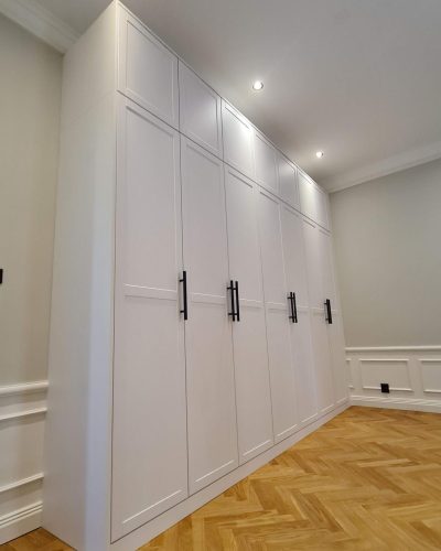 Platsbyggd vit garderob med svarta handtag, monterad i ett rum med fiskbensparkett och dekorativa vägglister. Infälld takbelysning ger ett elegant och modernt intryck. Renovering och finsnickeri i Stockholm.