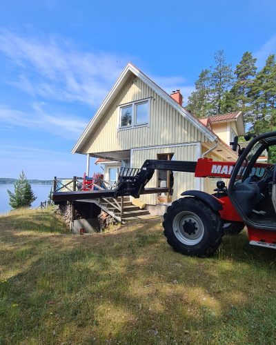Renovering av ett fritidshus vid vattnet i Stockholm. En röd teleskoplastare av märket Manitou står parkerad framför huset med sin lyftarm utsträckt mot den delvis rivna verandan. Det ljusgula trähuset har stora fönster och ligger omgivet av tallar, med utsikt över en sjö.