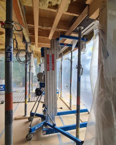 Pågående renovering i Stockholm med uppstöttat tak. Byggställningar och en blå materiallyft (SLA-20) används, samt stämp för att hålla upp konstruktionen. Isolering och elkablar syns i taket, och plastskydd täcker delar av arbetsområdet.