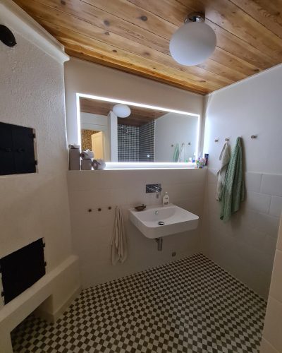 Nyrenoverat badrum med en kombination av rustik och modern stil. Rutigt klinkergolv, vitkaklade väggar och ett tak i naturligt trä skapar en varm känsla. Ett vägghängt handfat med kromad blandare är placerat under en stor spegel med inbyggd belysning. Till vänster syns en murad eldstad med svarta luckor. Gröna handdukar hänger på träkrokar. Renovering utförd i Stockholm.