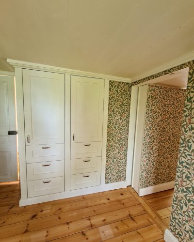 Nyrenoverat rum med platsbyggd vit garderob med lådor och mässingshandtag. Väggarna är klädda i en klassisk blommig tapet, och rummet har breda trägolv med varm ton. Renovering utförd i Stockholm.