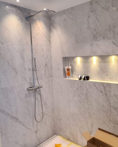 Nyrenoverad dusch med stora, marmorinspirerade kakelplattor och en inbyggd nisch med LED-belysning för förvaring. Takmonterad regndusch i krom ger en modern känsla. Dörren står öppen och en kartong syns på golvet, vilket tyder på pågående arbete. Badrumsrenovering i Stockholm.
