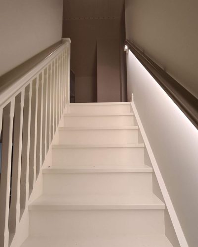 Nyrenoverad vit trappa med klassiskt räcke och inbyggd LED-belysning längs väggen. Taket är klätt i träpanel, och en infälld spotlight ger extra ljus. Renovering och totalentreprenad i Stockholm.