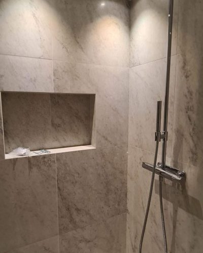 Nyrenoverad dusch med stora, ljusa marmorinspirerade kakelplattor och en inbyggd nisch för förvaring. Takmonterad regndusch i krom och infälld spotlightsbelysning skapar en modern känsla. Badrumsrenovering i Stockholm.