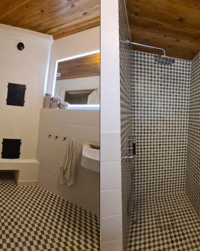 Nyrenoverat badrum med en unik kombination av klassisk och modern stil. Badrummet har rutigt klinkergolv, vitkaklade väggar och ett trätak som ger en varm känsla. En vit toalett och ett litet handfat är placerade bredvid en murad eldstad. Duschutrymmet har samma rutiga klinker på både golv och väggar samt en takdusch i krom. Renovering utförd i Stockholm.