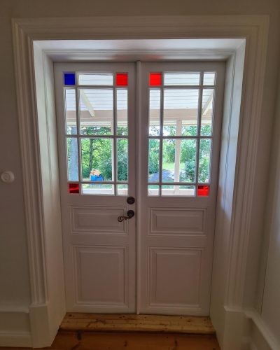 Nyrenoverad entré med vit dubbeldörr i klassisk stil, dekorerad med spröjsat glas och färgade detaljer i rött och blått. Trägolv och breda dörrfoder förstärker den traditionella känslan. Renovering utförd i Stockholm.