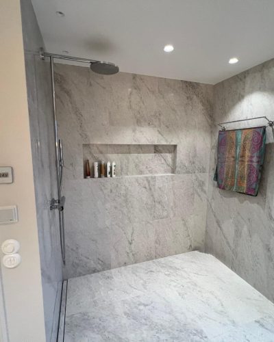 Elegant nyrenoverat badrum med dusch i natursten och infälld hylla för förvaring. Takdusch i krom och inbyggd belysning skapar en modern känsla. En färgglad handduk hänger på en kromad handdukshållare. Renovering utförd i Stockholm.